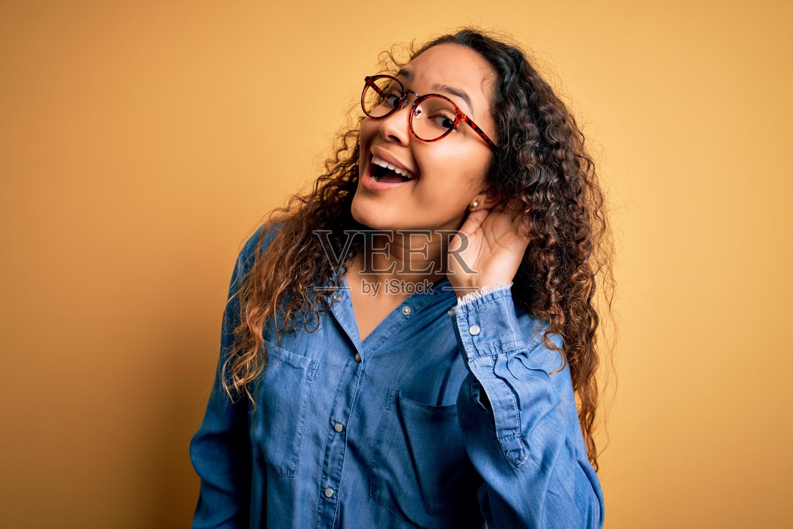 穿着休闲牛仔衬衫、戴着眼镜的卷发美女，倾听谣言或八卦，耳聋概念照片摄影图片