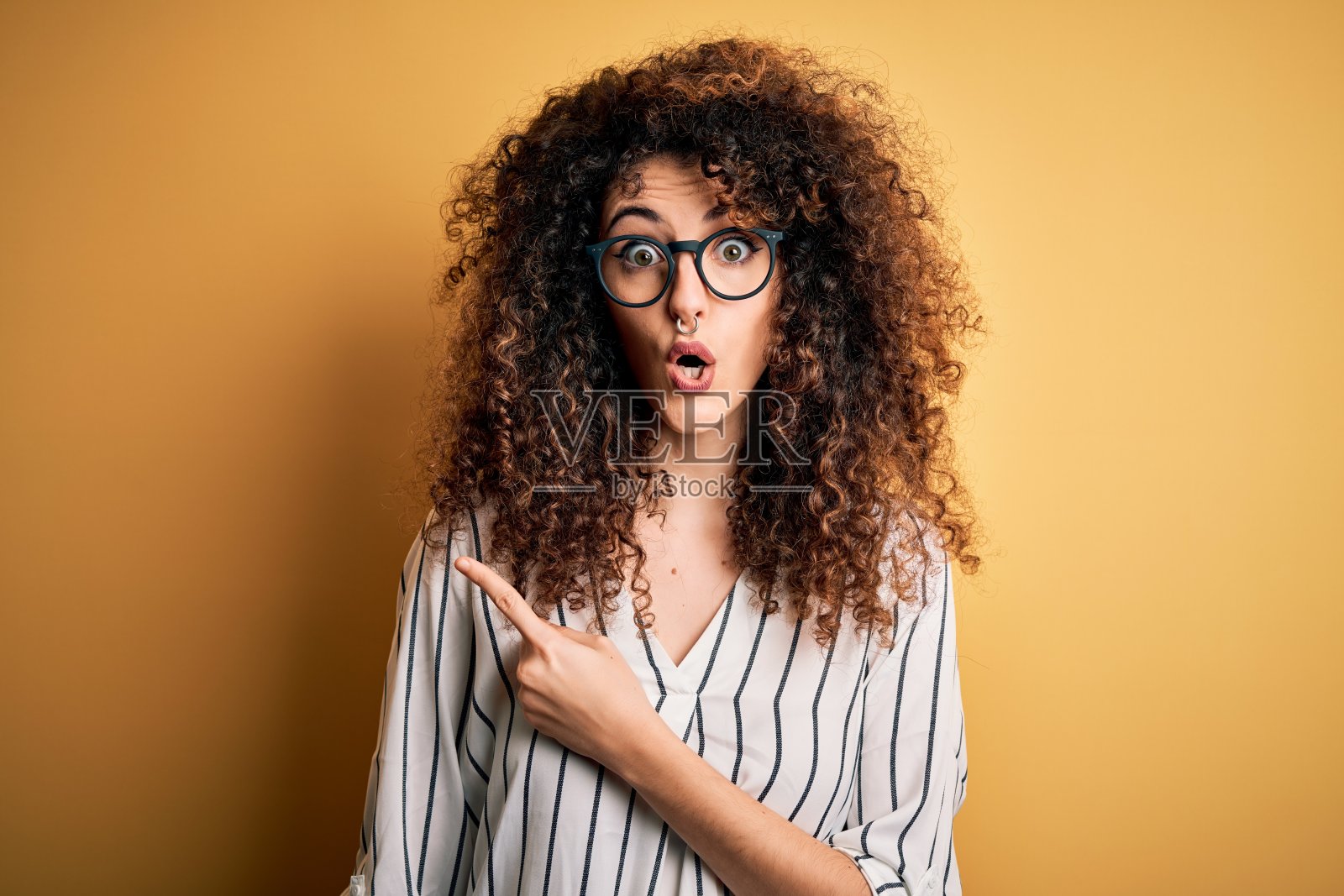 年轻漂亮的女子，一头卷发，穿条纹衬衫和眼镜惊讶地用手指向一边，张大嘴巴惊讶的表情。照片摄影图片