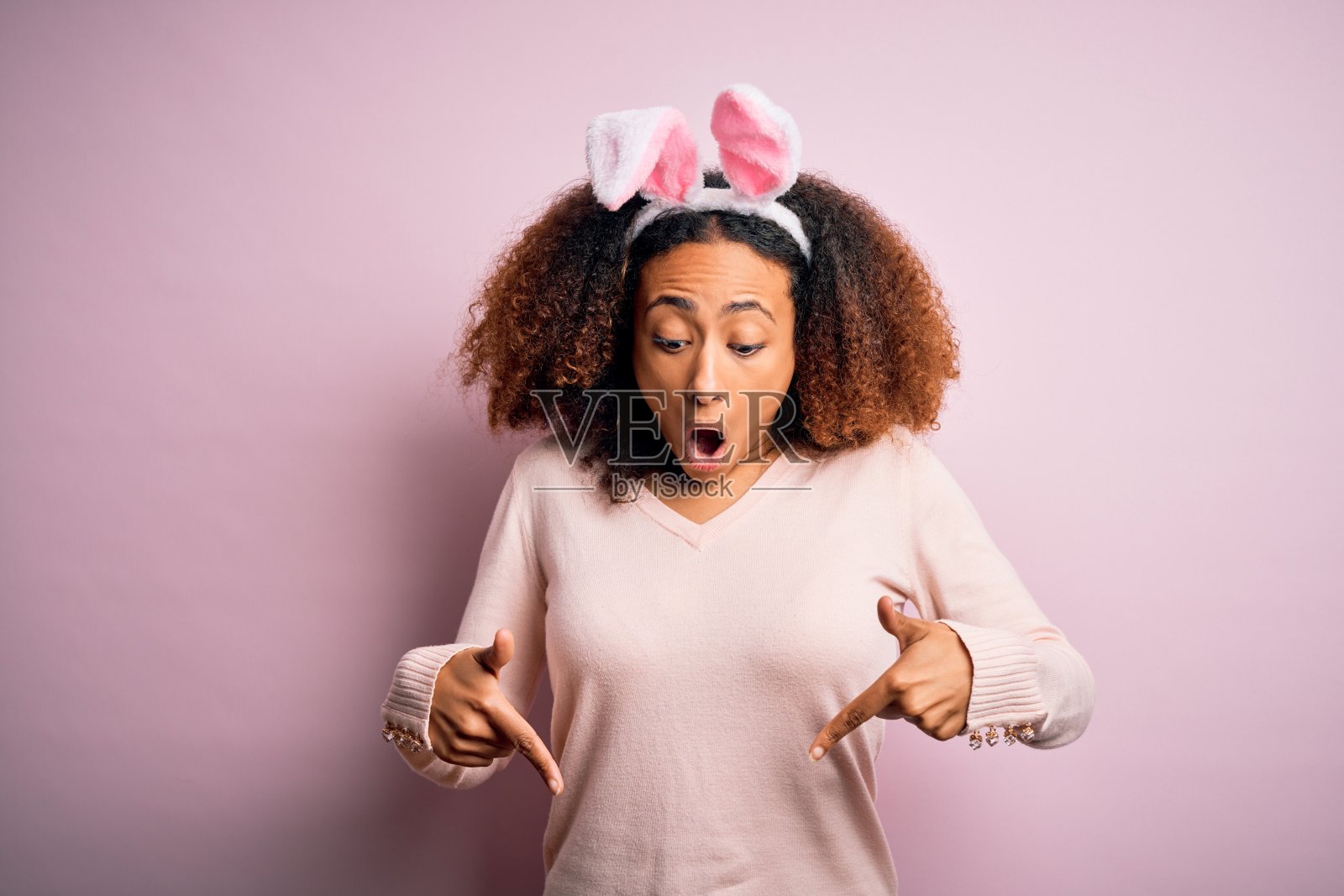 年轻的非裔美国女性在粉色背景下戴着兔子耳朵，用手指指向下方，展示广告，面带惊讶和张着嘴巴照片摄影图片