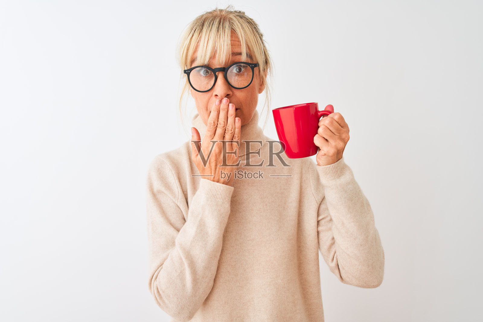 戴着眼镜的中年妇女在白色背景前喝着红色杯子里的咖啡，用手捂住嘴巴，因犯错而感到震惊和羞愧，表达了恐惧，在沉默中感到害怕，秘密的概念照片摄影图片