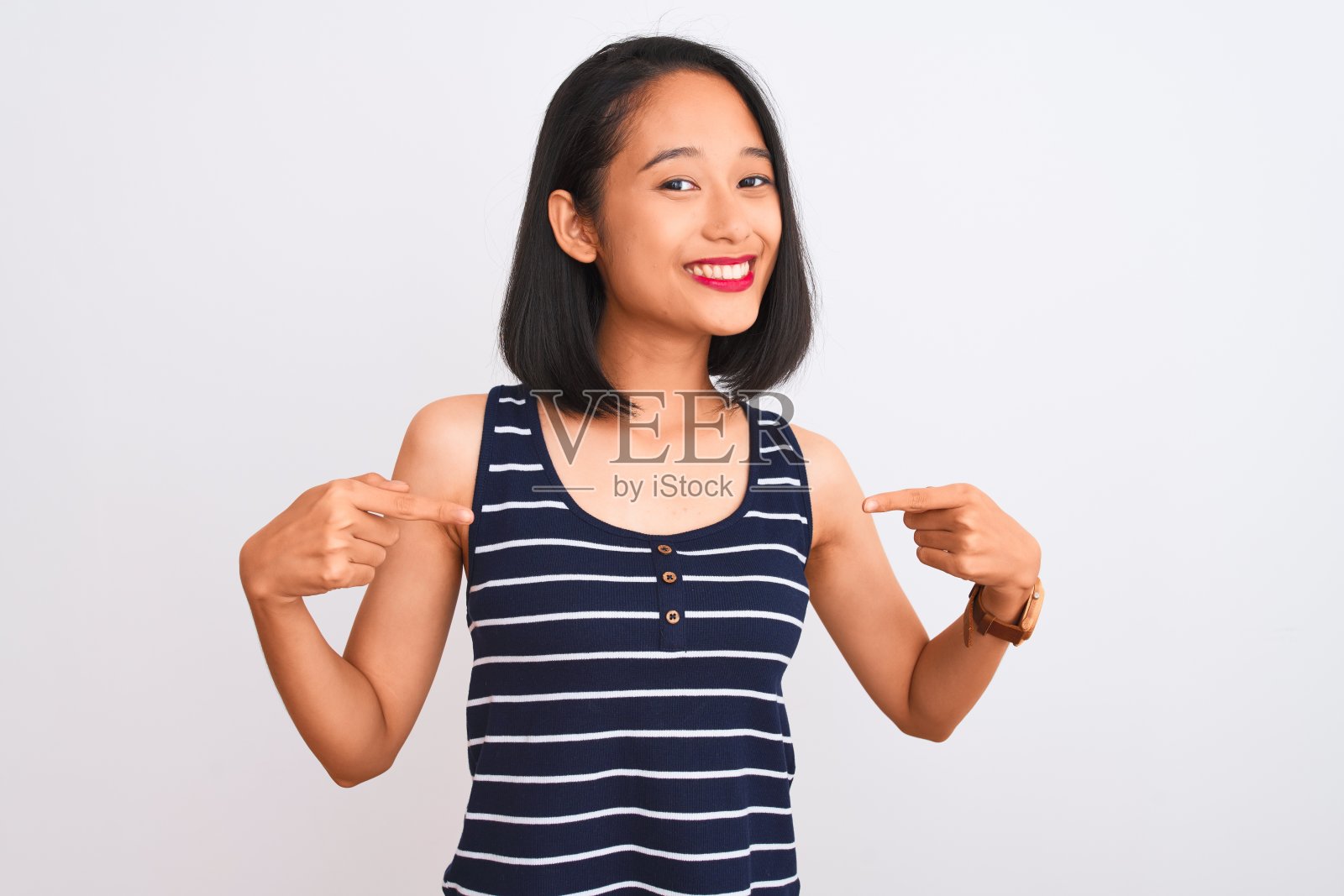 年轻的中国女子穿着条纹t恤站在孤立的白色背景看起来自信，脸上带着微笑，用手指指着自己骄傲和幸福。照片摄影图片