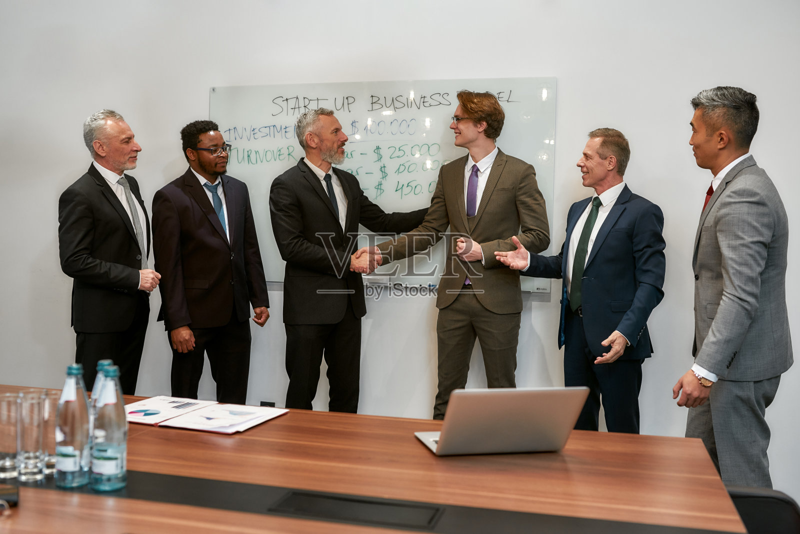 交易：两位穿着经典商务装的快乐商人在现代办公室与团队会面并握手照片摄影图片