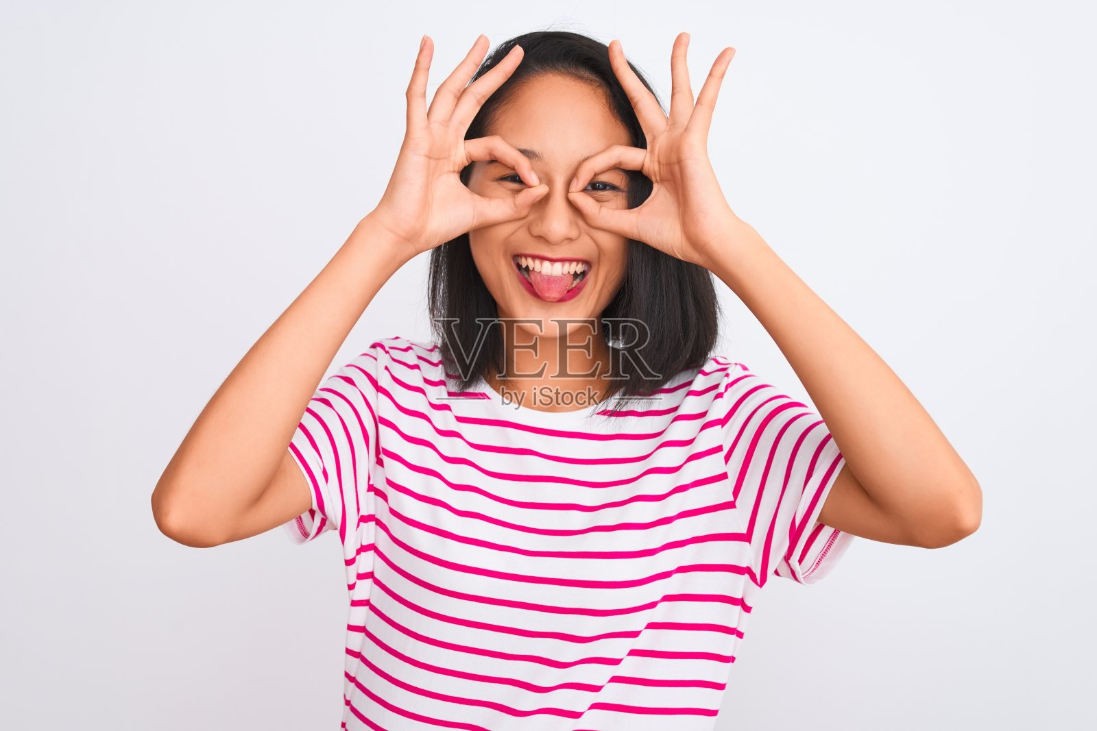 一个年轻的中国女人穿着条纹t恤站在孤立的白色背景下做着不错的手势，就像望远镜伸出舌头，眼睛透过手指看。疯狂的表情。照片摄影图片