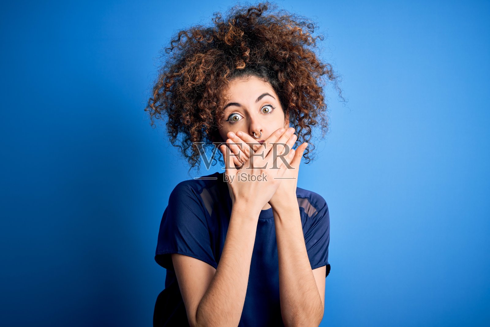 年轻美丽的女子与卷发和穿休闲蓝色t恤震惊掩盖嘴与手的错误。秘密的概念。照片摄影图片
