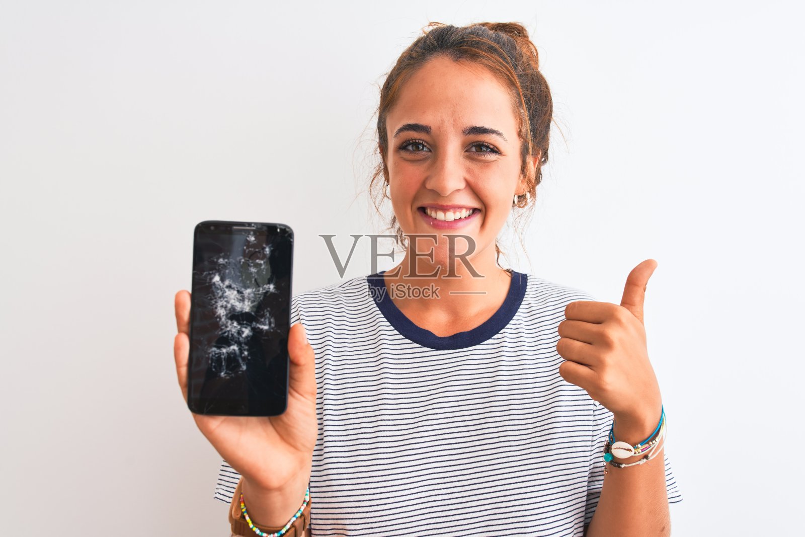 年轻的红发女子拿着破碎的智能手机在孤立的背景下，开心地笑着做ok的手势，用手指竖起大拇指，优秀的手势照片摄影图片