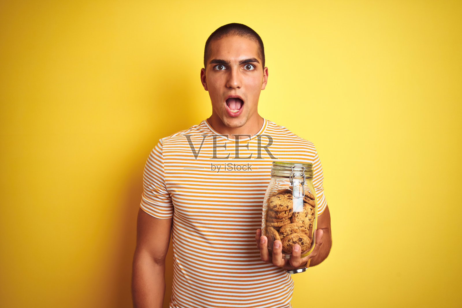 年轻英俊的男子拿着一罐饼干越过黄色孤立的背景吓得目瞪口呆，脸上带着惊讶、害怕和兴奋的恐惧表情照片摄影图片