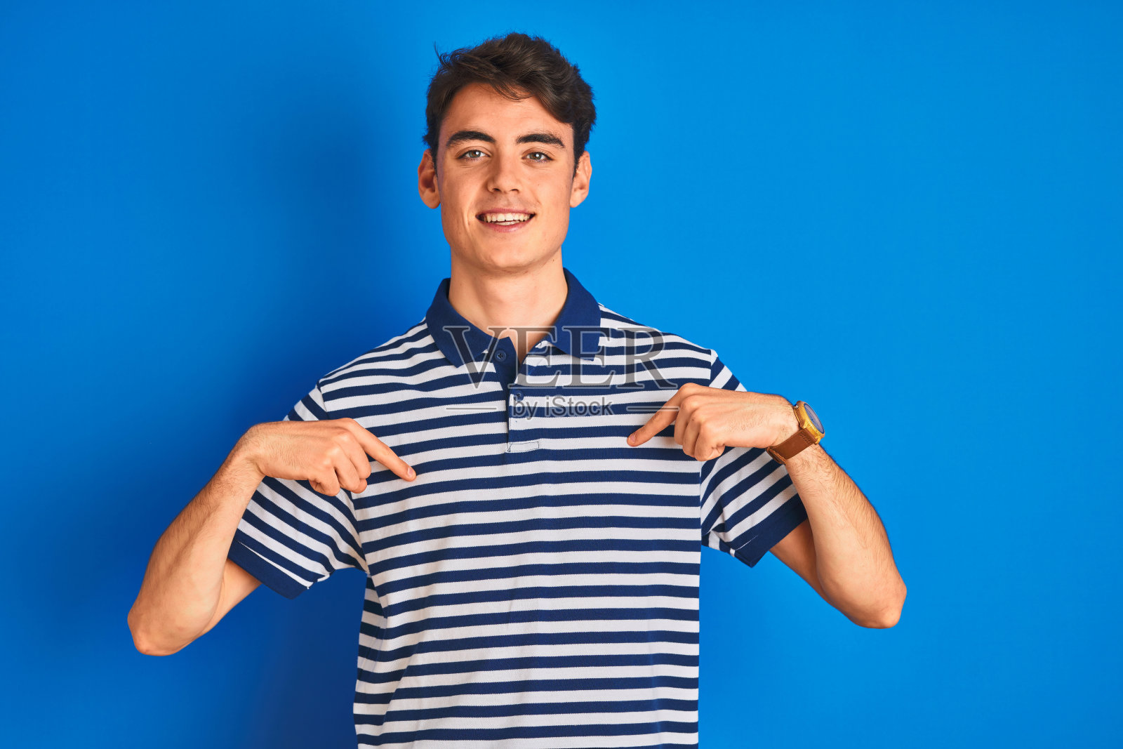 少年穿着休闲T恤站在蓝色背景前，面带微笑，自信地用手指指向自己，感到自豪和快乐照片摄影图片