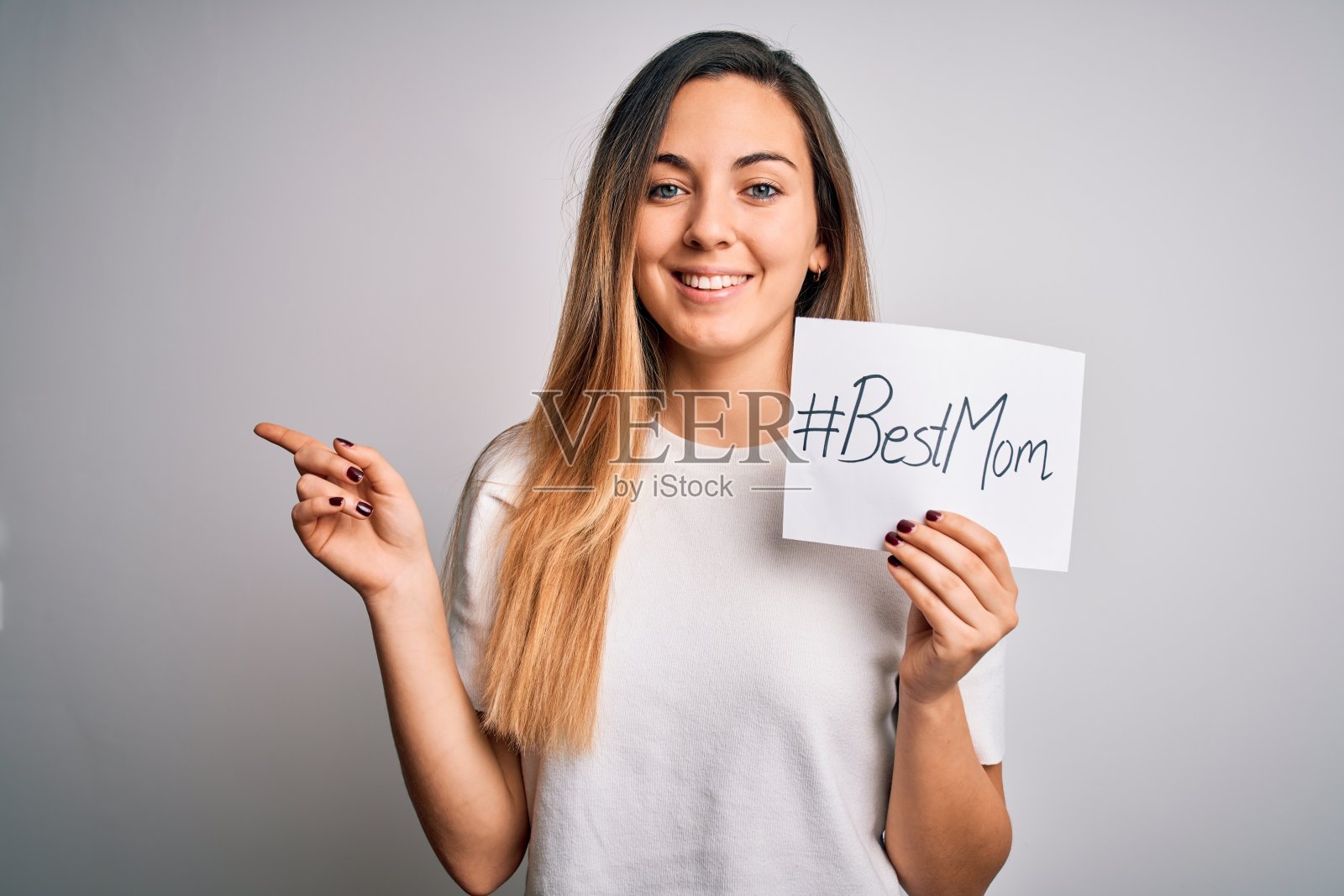 年轻漂亮的女人拿着纸和最好的妈妈短信庆祝母亲节非常快乐，用手和手指指着一边照片摄影图片