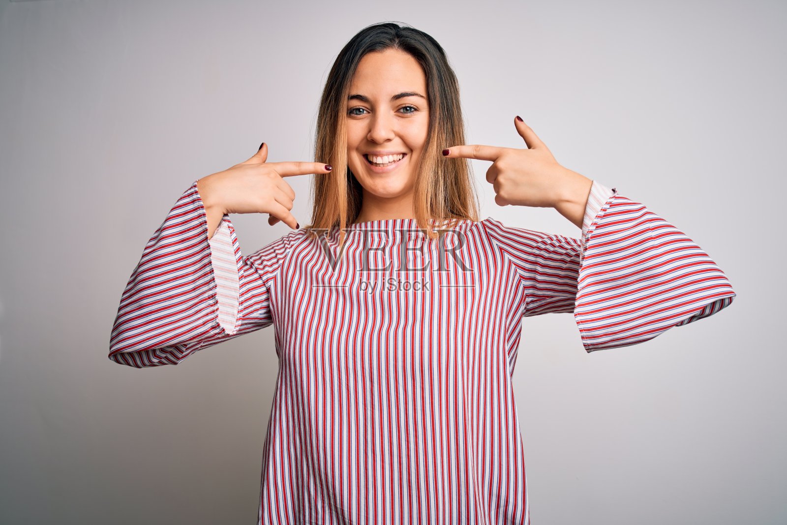 年轻美丽的金发女人与蓝色的眼睛穿针t恤在白色的背景微笑愉快显示和指向手指，牙齿和嘴。牙齿健康的概念。照片摄影图片