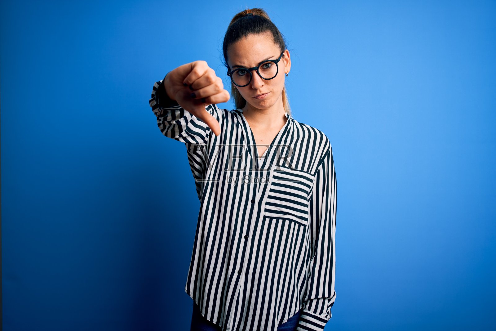 美丽的金发女人，蓝色眼睛，穿着条纹衬衫和眼镜在蓝色的背景看起来不开心和生气，显示拒绝和负面的拇指向下的手势。糟糕的表达。照片摄影图片