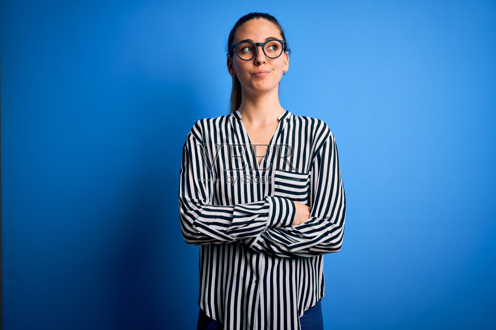 美丽的金发女人与蓝色眼睛穿着条纹衬衫和眼镜在蓝色的背景微笑看向一边和凝视远离思考。照片摄影图片