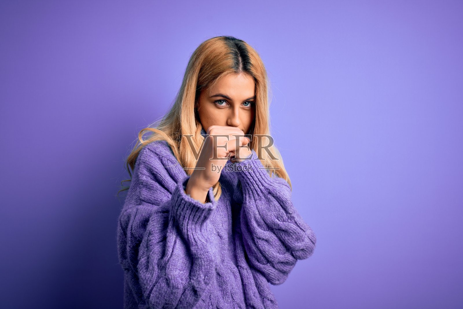 年轻美丽的金发女子穿着休闲高领毛衣越过紫色的背景准备打架用拳头防御手势，愤怒和不安的脸，害怕问题照片摄影图片