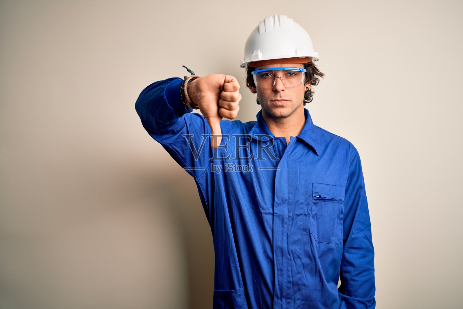 年轻的建筑工人穿着制服和安全帽，在孤立的白色背景下看起来不高兴和生气，用拇指向下的手势表示拒绝和否定。糟糕的表达。照片摄影图片