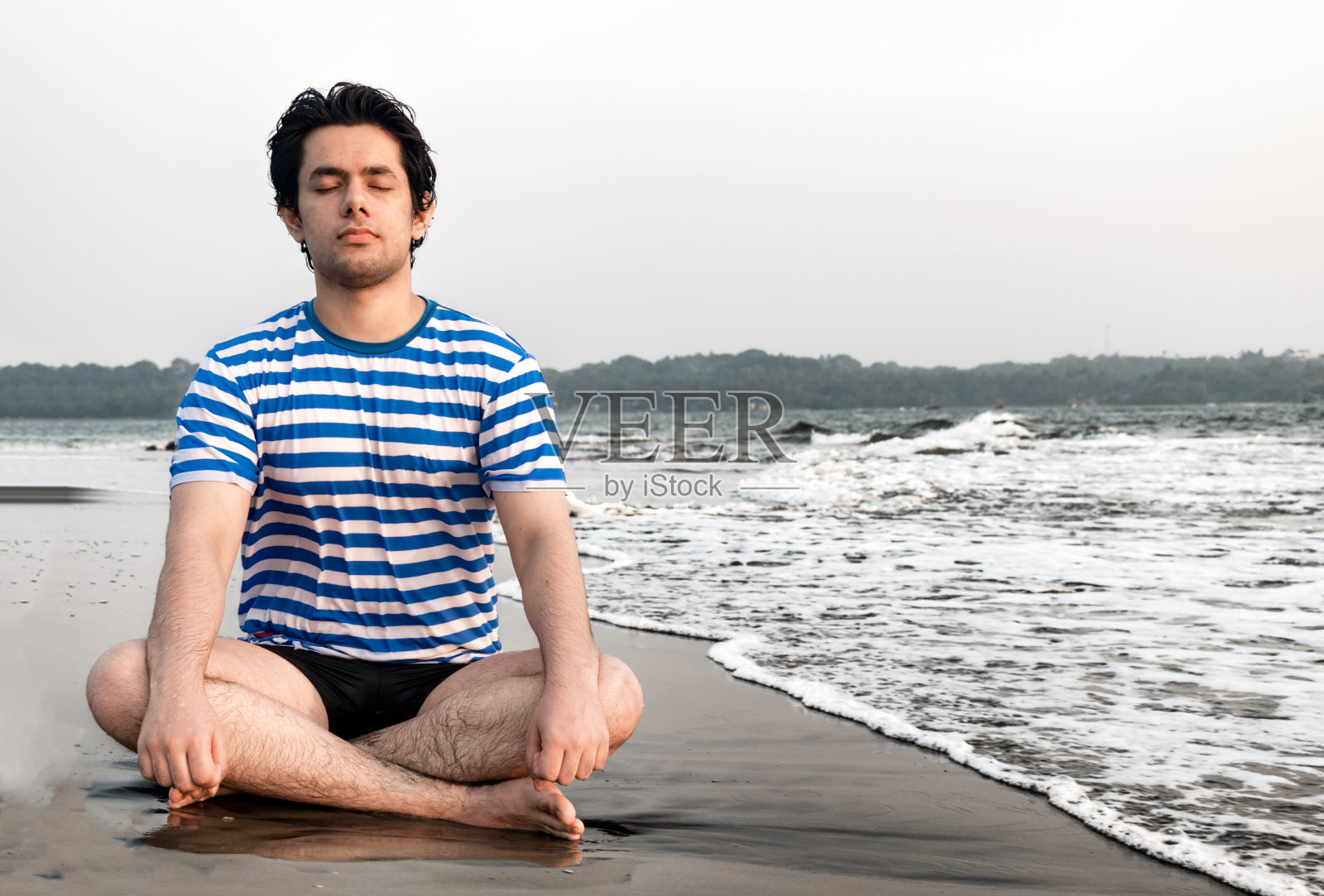 20岁出头的英俊男孩在海滩上以莲花姿势冥想，以获得身心健康照片摄影图片