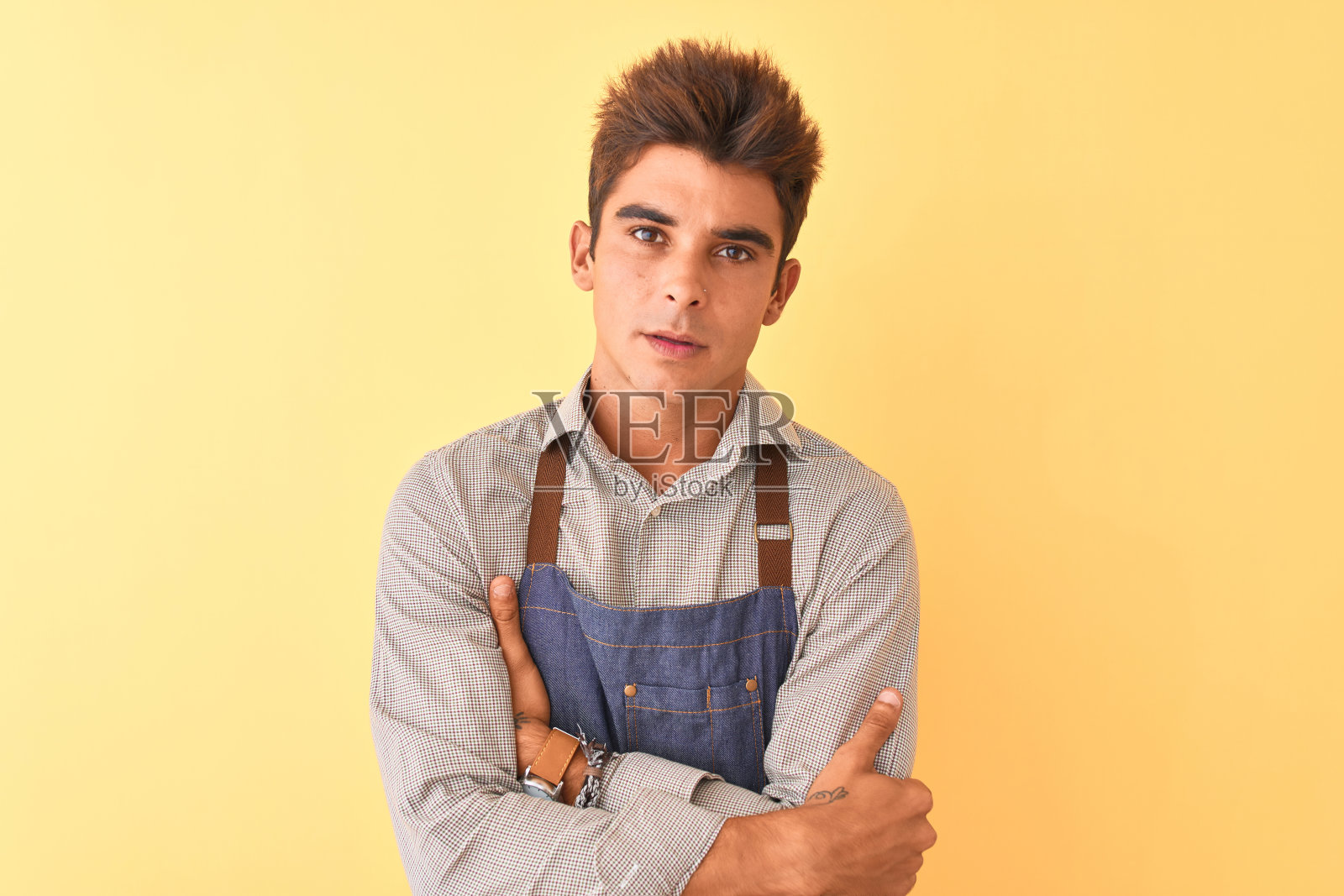 年轻英俊的员工男子穿着围裙站在孤立的黄色背景怀疑和紧张，不赞成的表情脸上交叉双臂。消极的人。照片摄影图片