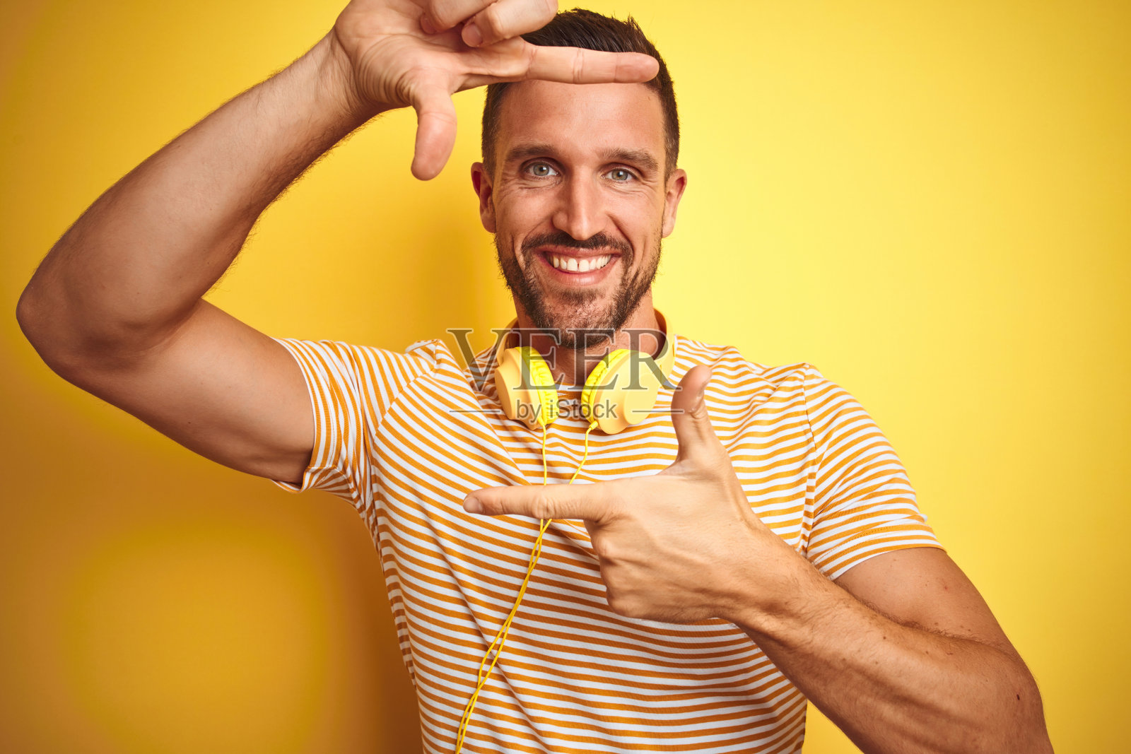 戴着耳机听音乐的年轻英俊男子在黄色背景前微笑，用手和手指比出取景框，表达创意和摄影概念照片摄影图片