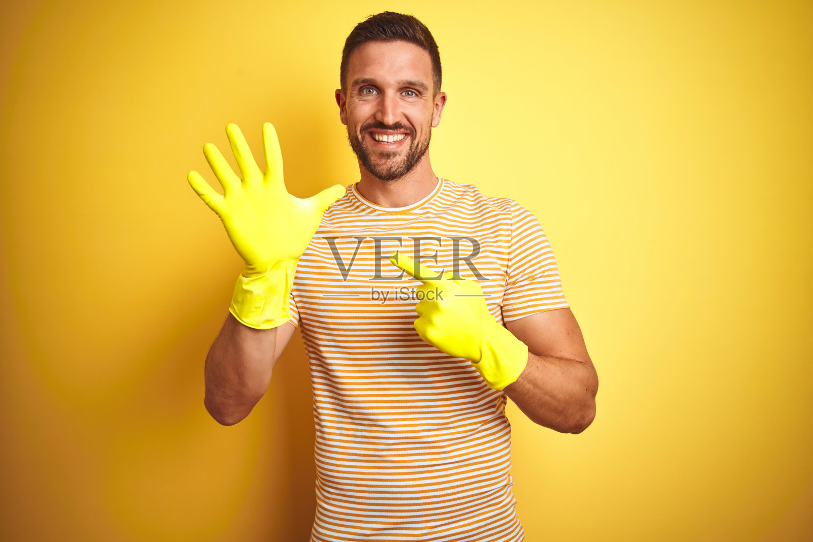 戴着家务清洁手套的年轻帅气男子在黄色背景前高兴地用手指指着照片摄影图片