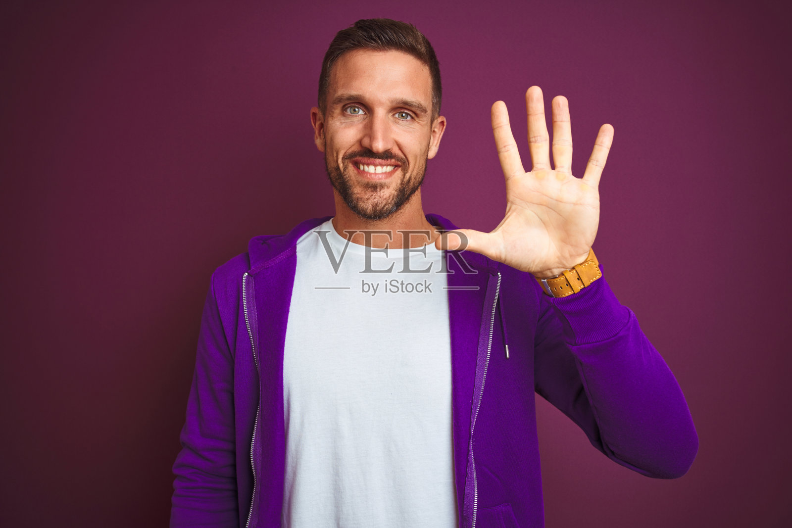 穿着休闲运动衫的年轻健身男人，在紫色背景前自信地笑着，用手指指向上方，比出数字五照片摄影图片