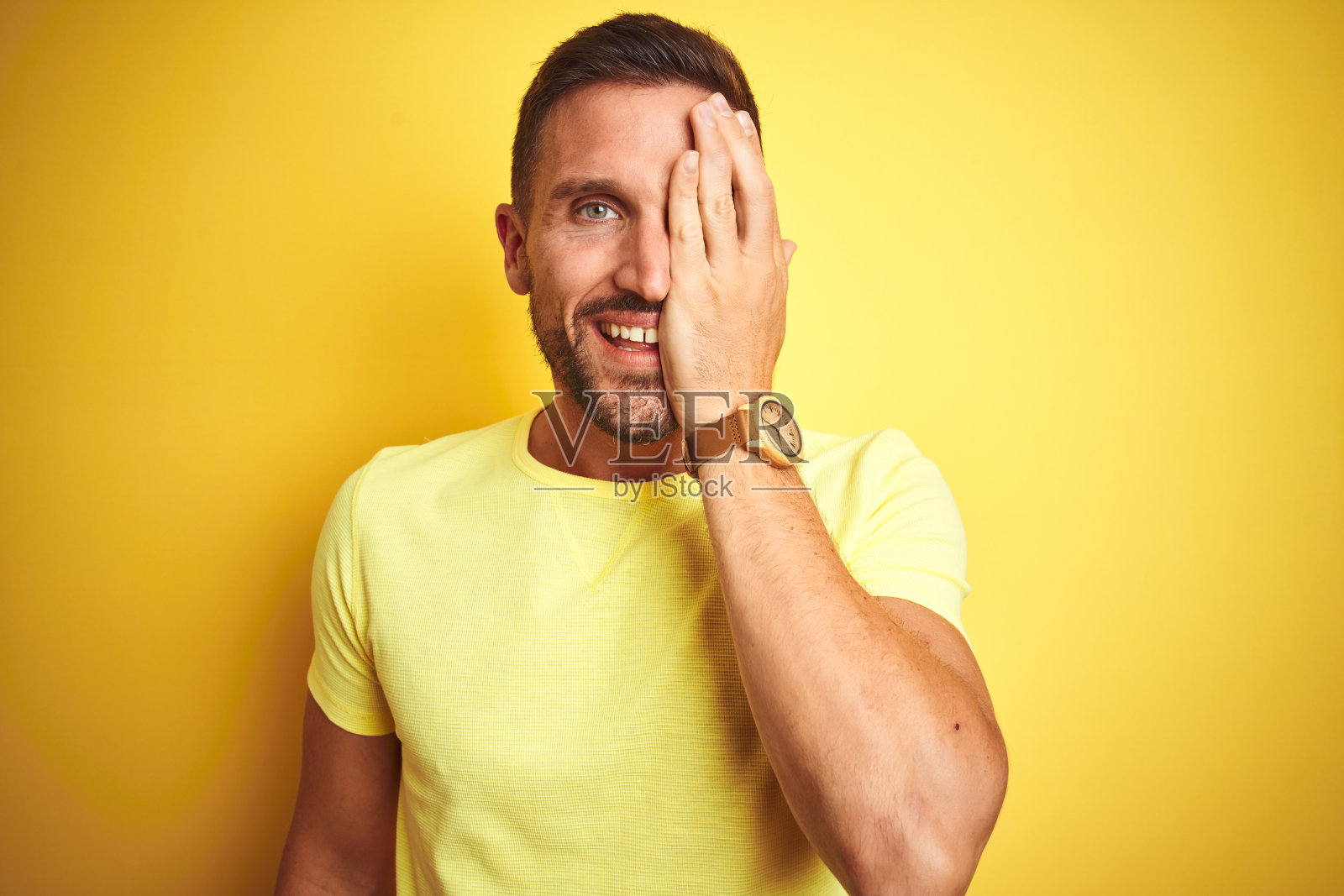 年轻帅气的男人穿着休闲黄色T恤在黄色背景前，用手遮住一只眼睛，脸上带着自信的微笑和惊讶的表情照片摄影图片