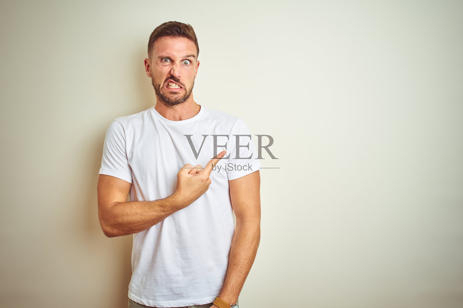 穿着休闲白色T恤的年轻英俊男人，用食指指向一侧，担心和紧张，表情关注和惊讶照片摄影图片