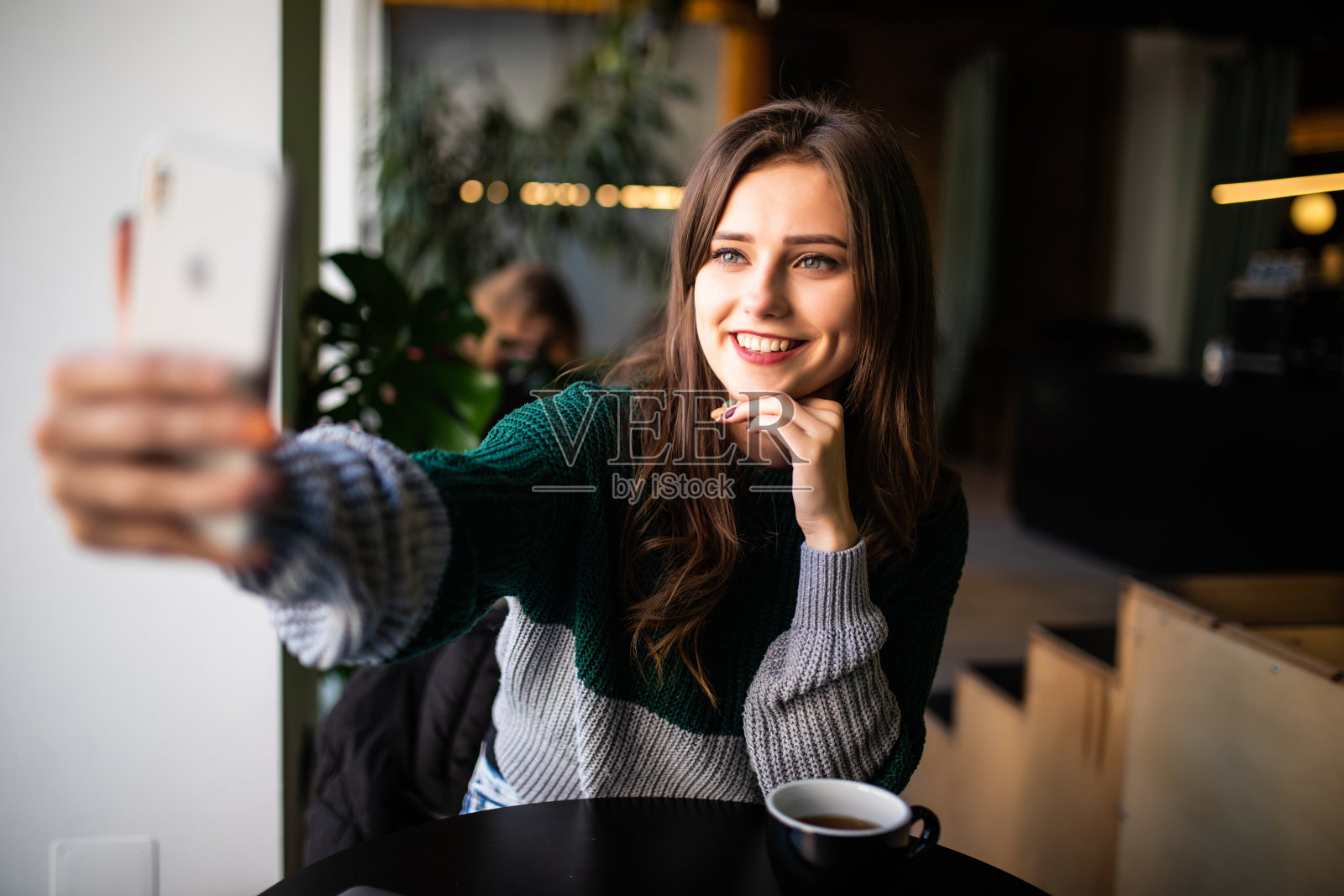 一名年轻女子坐在桌旁，手里拿着一杯热咖啡，用智能手机自拍照片摄影图片