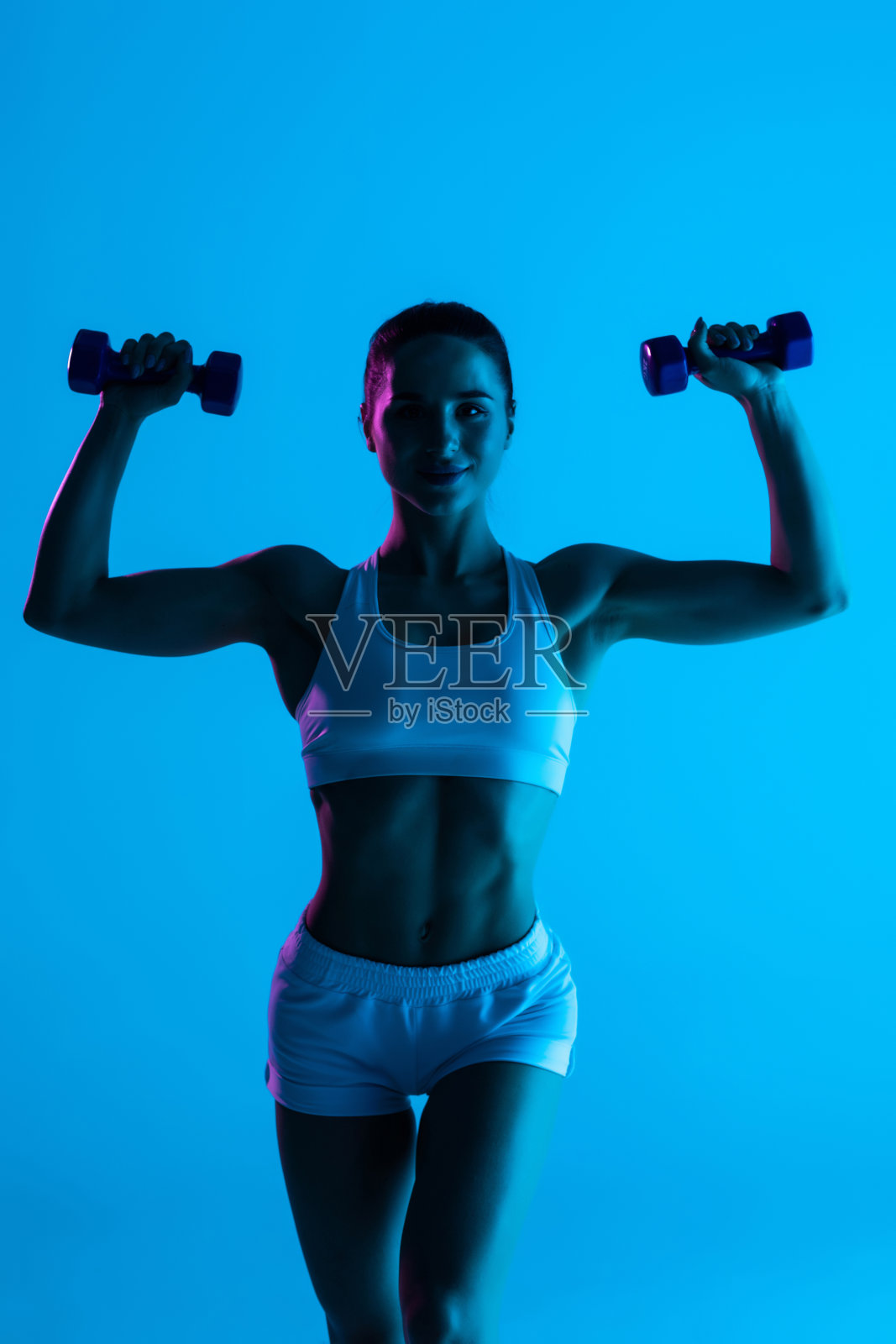 健身的年轻女子用哑铃在蓝光背景下锻炼照片摄影图片