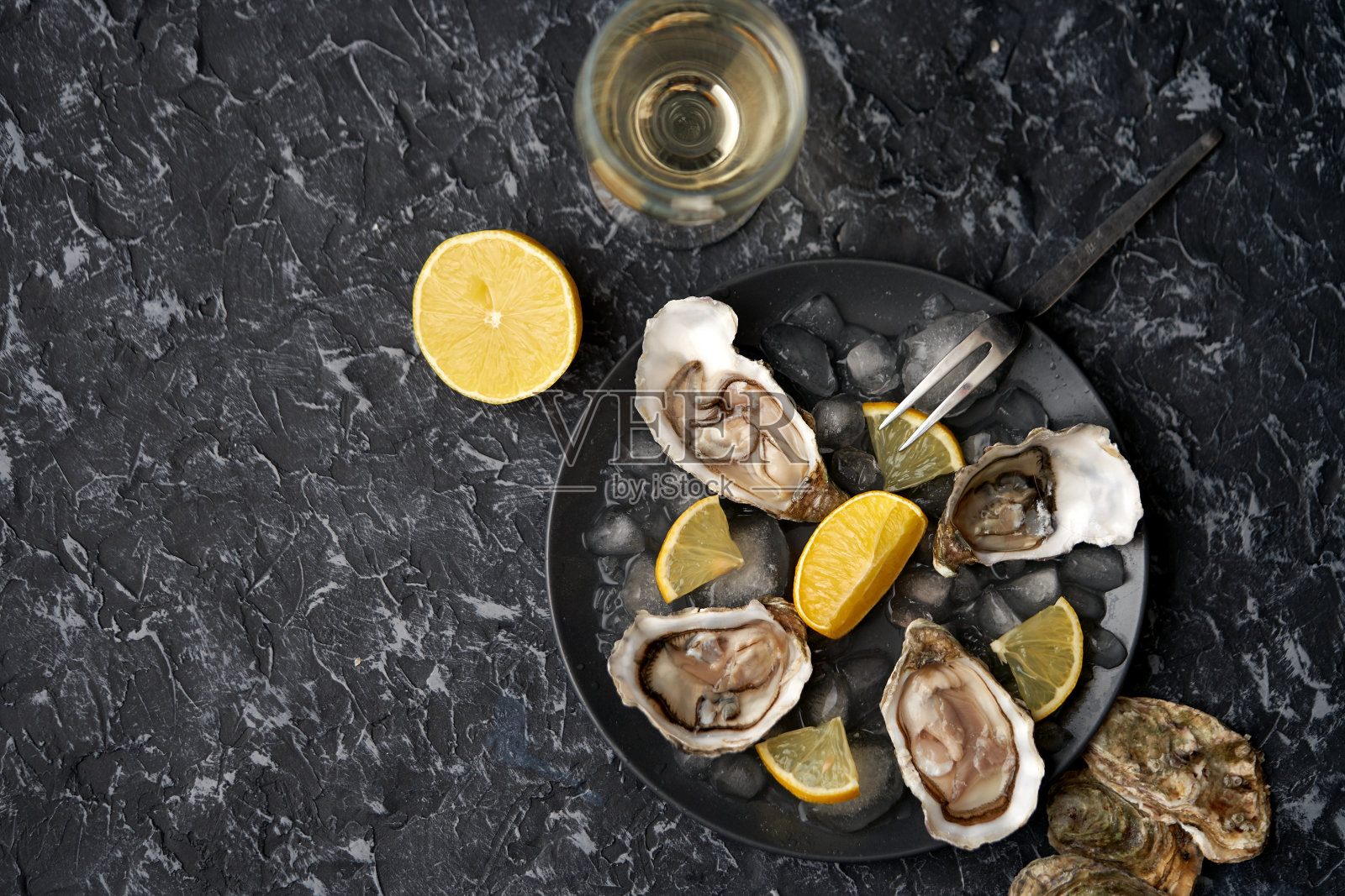 用柠檬角打开牡蛎，将碎冰放在黑色的盘子里，背景是黑色的石头和牡蛎壳，配上一杯香槟和牡蛎叉子。俯视图，处有题字，海鲜，豪华健康食品照片摄影图片