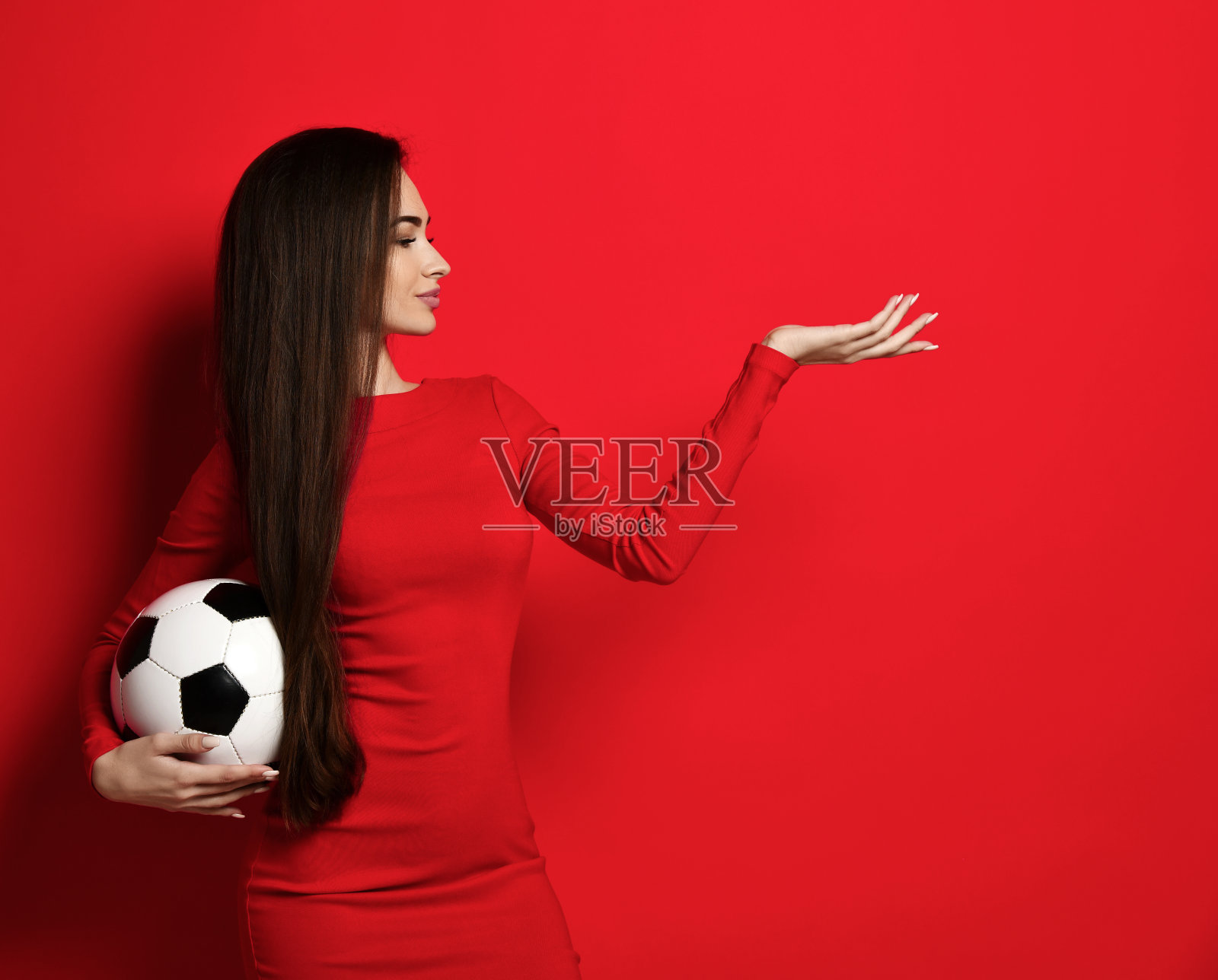 微笑的棕发女人穿着红色紧身连衣裙，手拿足球指着红色背景上的开放手掌照片摄影图片