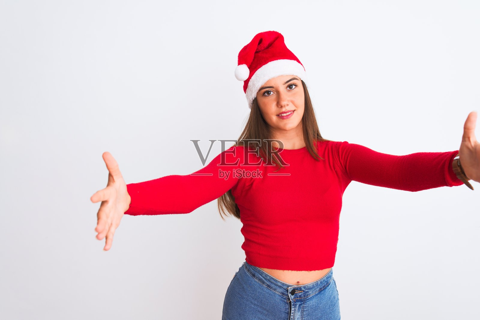 年轻美丽的女孩戴着圣诞老人的帽子站在孤立的白色背景看着镜头微笑着张开双臂拥抱。快乐的表情拥抱幸福。照片摄影图片