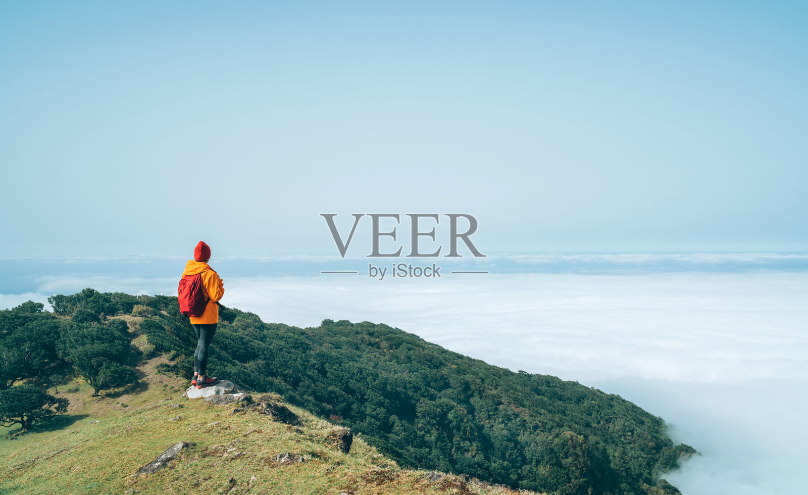 2月底，在葡萄牙马德拉岛，年轻的女背包客穿着橙色防水夹克，沿着云层上方的山徒步旅行。积极的人环游世界的概念。照片摄影图片