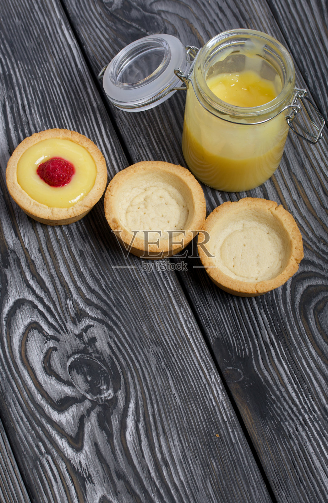 馅饼配柠檬布丁。装饰着覆盆子。他们躺在刷过的黑色松木板上。有一个开着的罐子，里面装着柠檬库德。旁边是没有馅料的馅饼。照片摄影图片