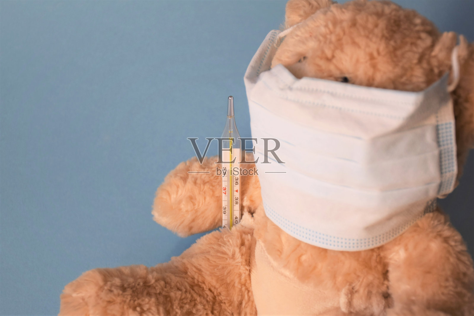 白色医用口罩上的泰迪熊幼崽坐在蓝色背景上，带温度计，概念COVID-19照片摄影图片