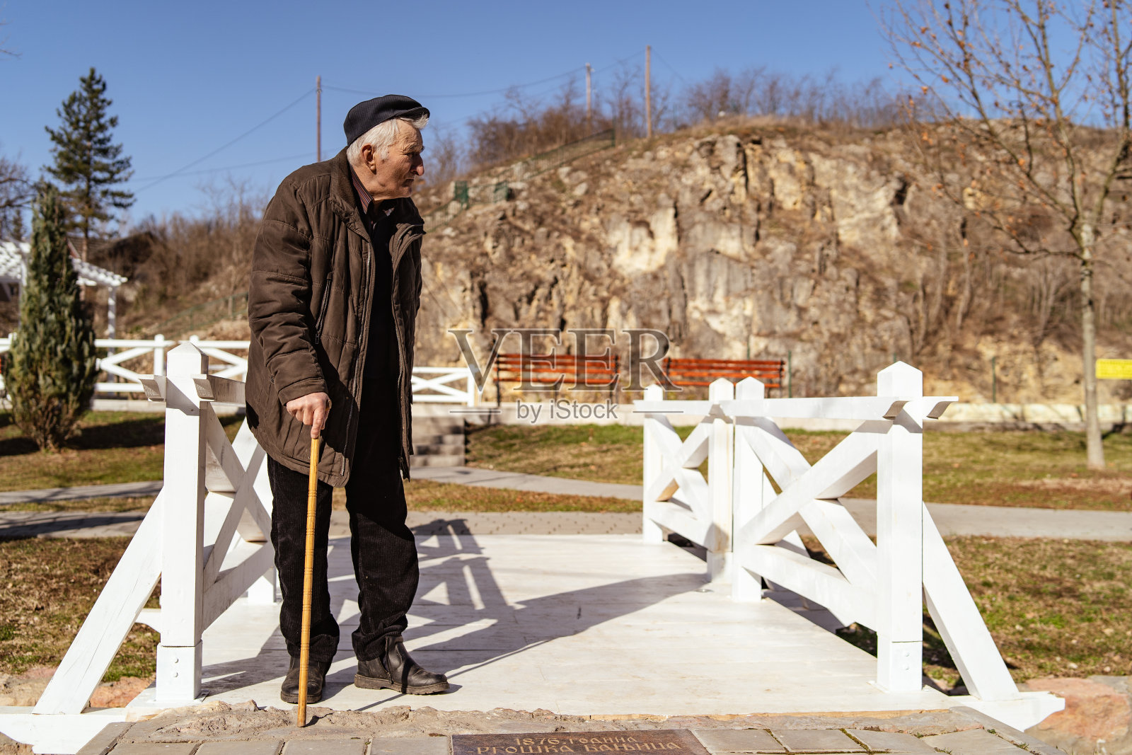 老爷爷拄着拐杖站在阳光明媚的白桥上或春日里，边看边穿着外套外套和帽子照片摄影图片