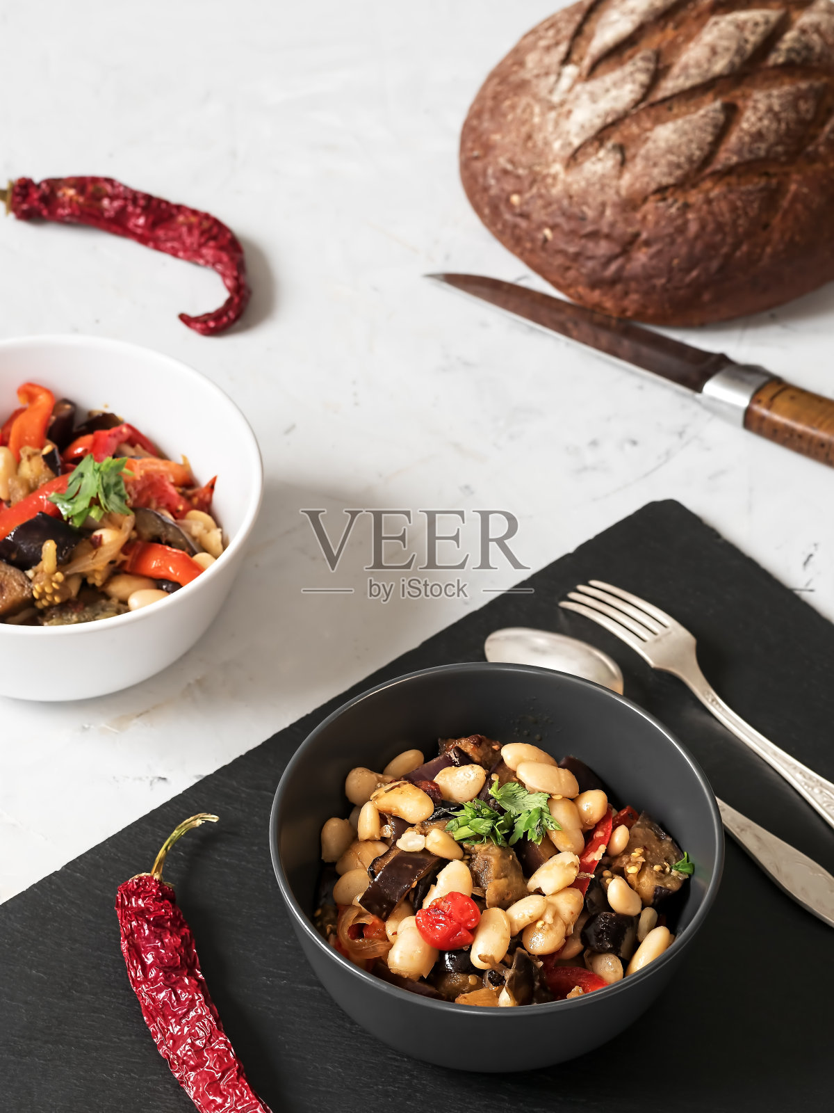 餐桌上的菜肴有炖茄子、豆子和红辣椒，再用一小枝香菜作装饰。黑白板，黑白背景。特写镜头。素食健康食品。照片摄影图片