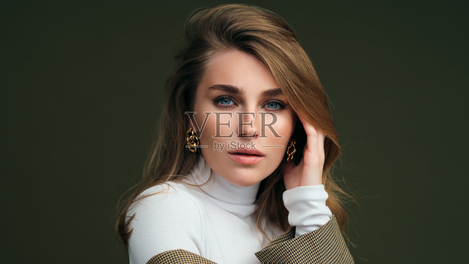 一名年轻的金发女子身穿白色高领毛衣和一件男士夹克，在画室里以橄榄绿为背景拍照。照片摄影图片