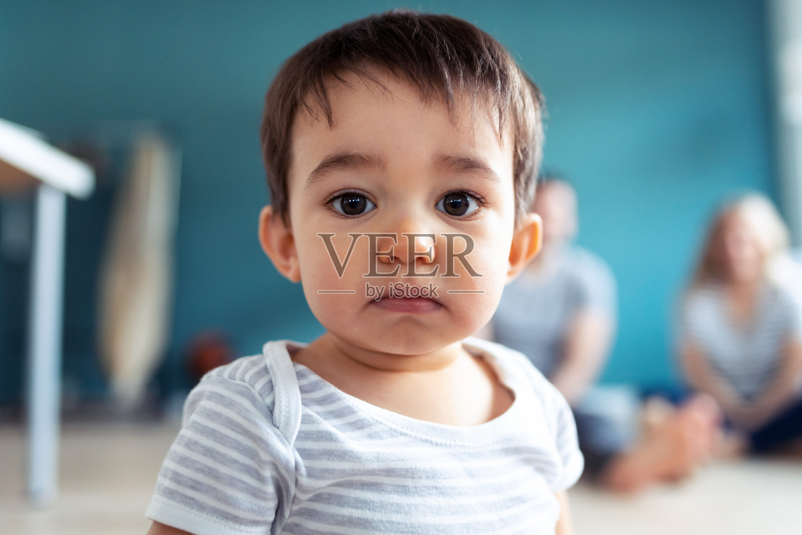 宝宝坐在家里的地板上看镜头。在背后，他的父母坐在一边看着他。照片摄影图片