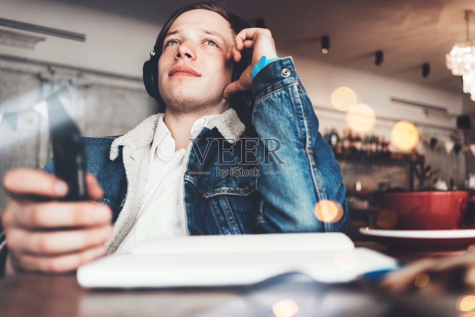 年轻快乐的男人在咖啡馆用耳机听音乐。手机，文书工作和一杯咖啡。聪明的休闲服装照片摄影图片