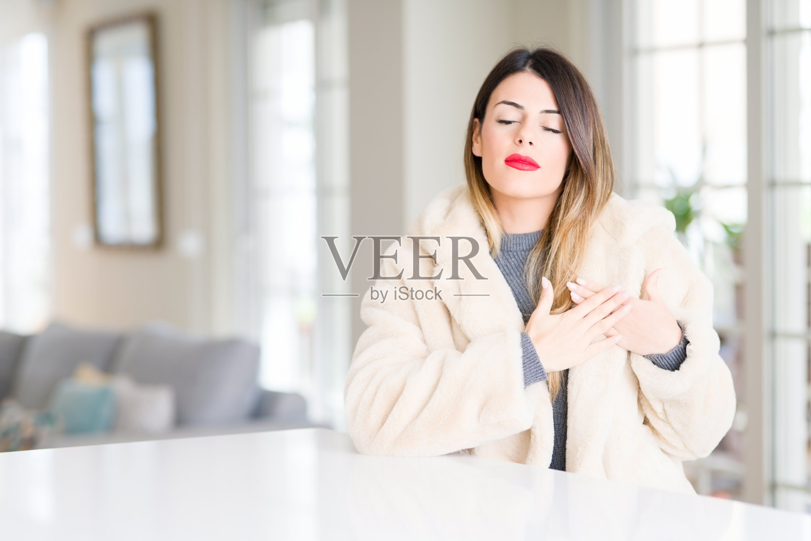 年轻漂亮的女人穿着冬天皮大衣在家微笑，双手放在胸前，闭上眼睛，脸上有感激的手势。健康的概念。照片摄影图片