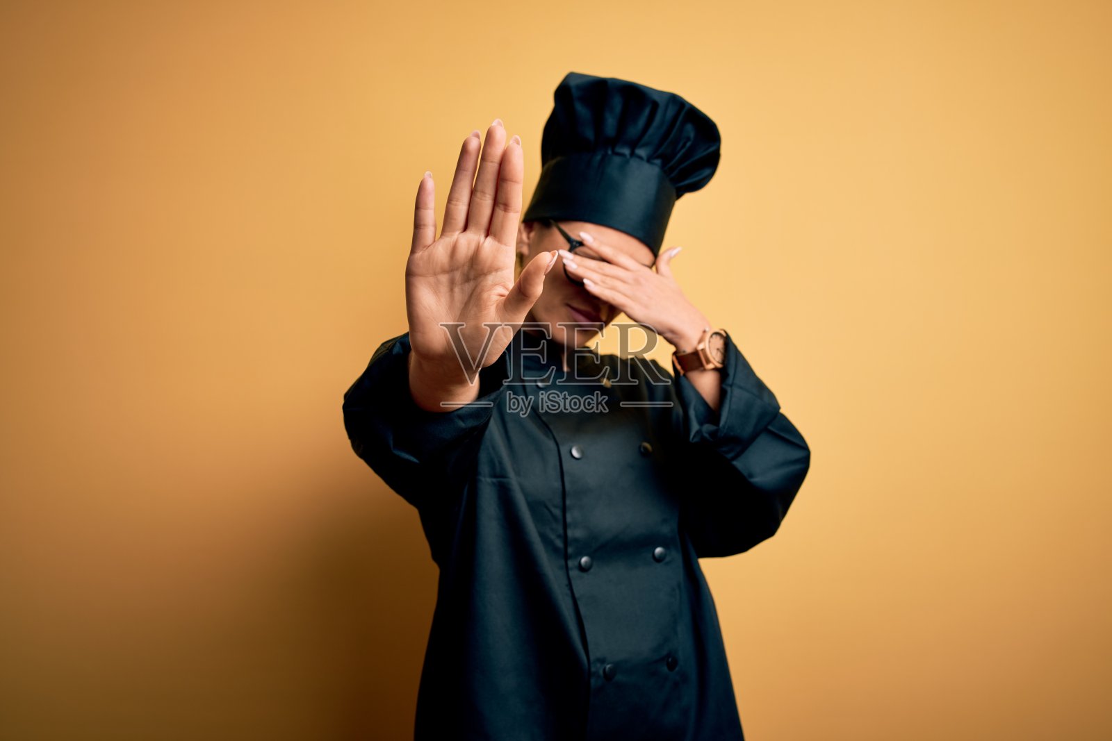 年轻美丽的褐发女厨师穿着炊具制服和帽子在黄色的背景覆盖眼睛与手和做停止手势与悲伤和恐惧的表情。尴尬和消极的概念。照片摄影图片