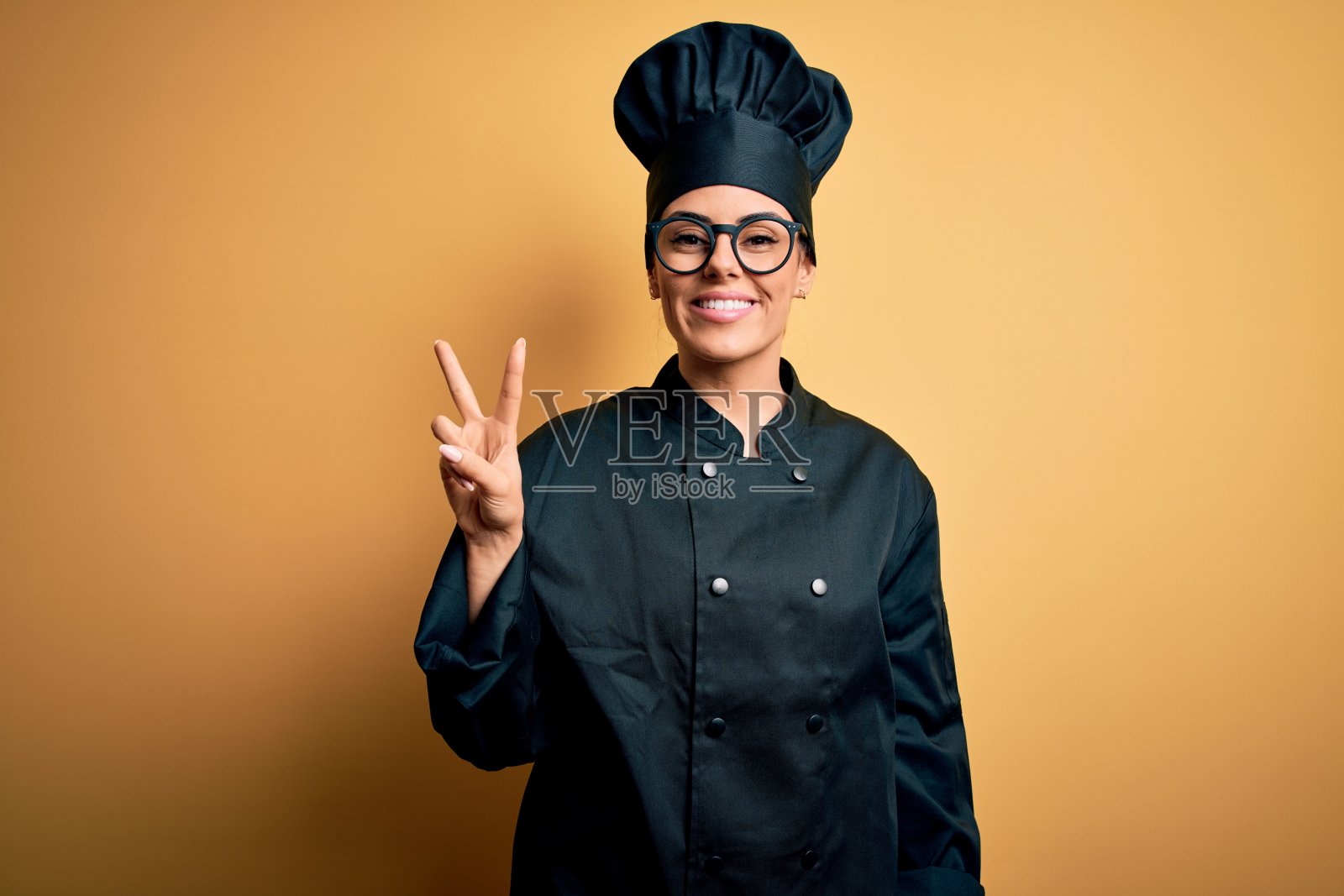 年轻美丽的褐发女厨师穿着炊具制服和帽子在黄色的背景显示和指向2号手指，同时微笑着自信和快乐。照片摄影图片