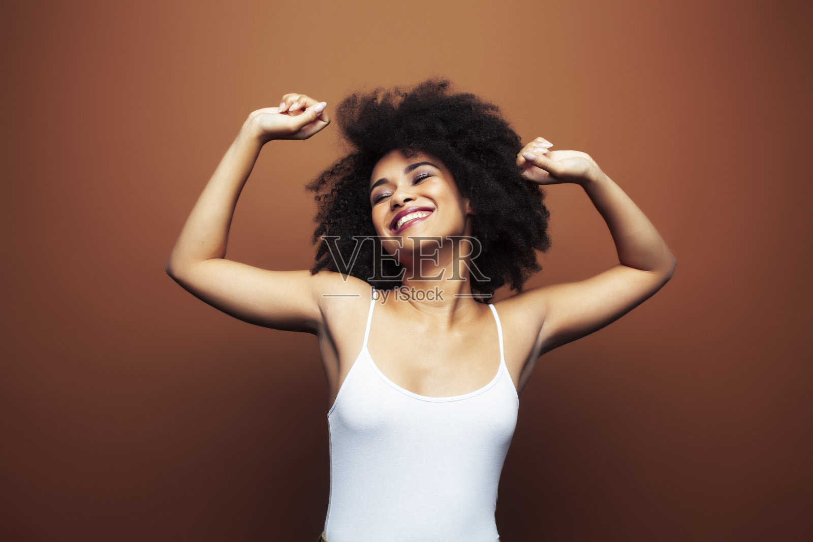 漂亮的年轻的非洲裔美国女人与卷曲的头发摆出愉快的手势在棕色的背景，生活方式的人的概念照片摄影图片