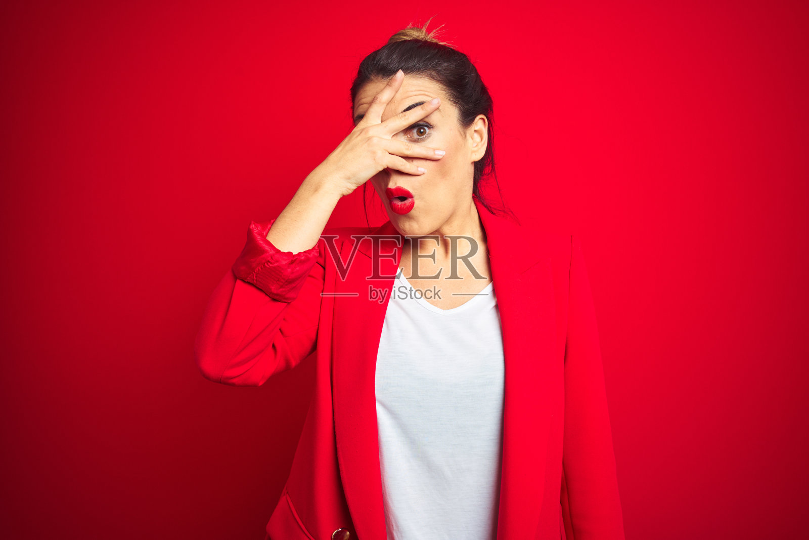 年轻美丽的女商人站在红色孤立的背景中窥视在震惊覆盖的脸和眼睛用手，透过手指看着尴尬的表情。照片摄影图片