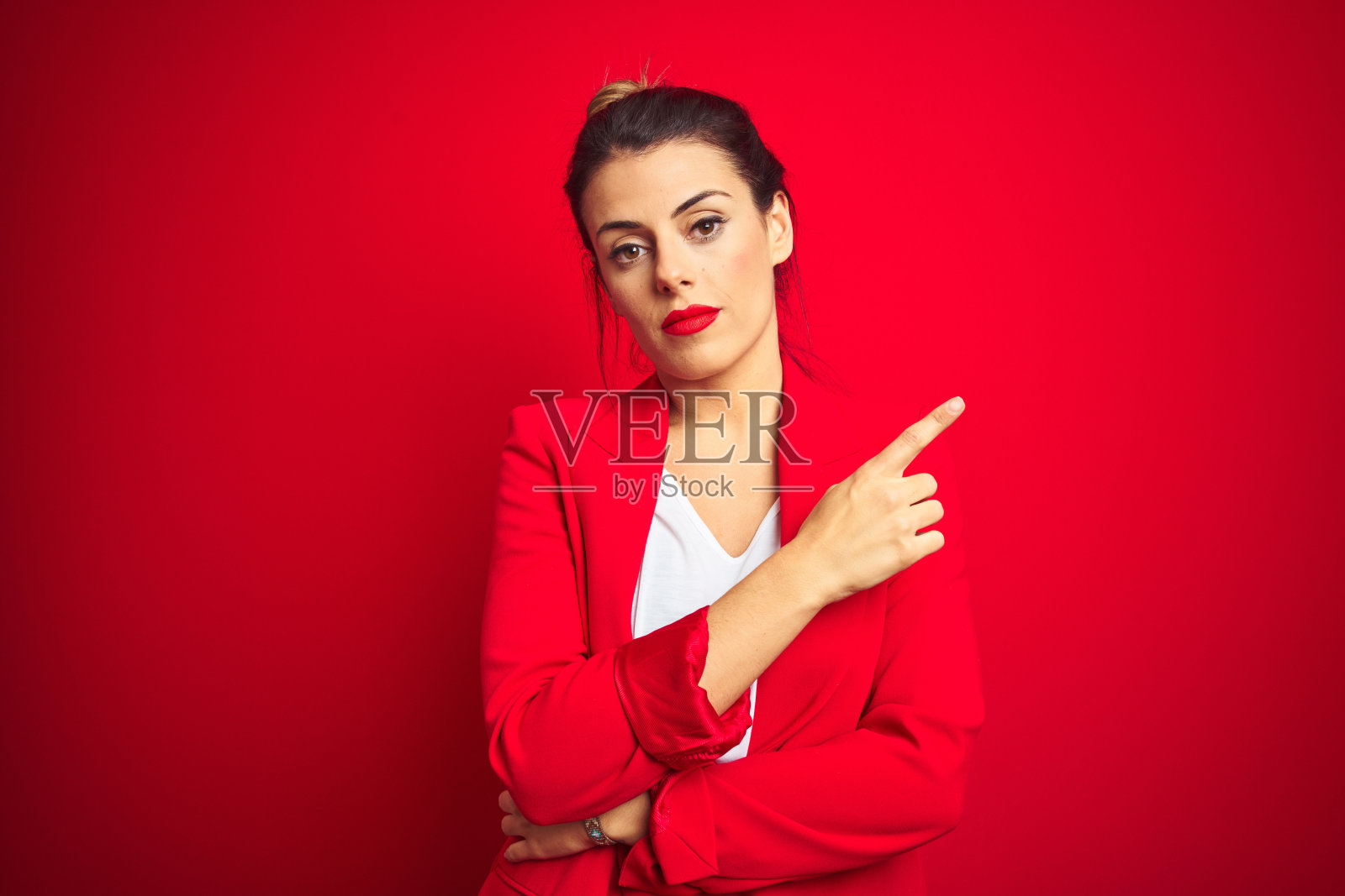 年轻美丽的女商人站在红色孤立的背景，手指指向旁边显示的广告，严肃而冷静的脸照片摄影图片