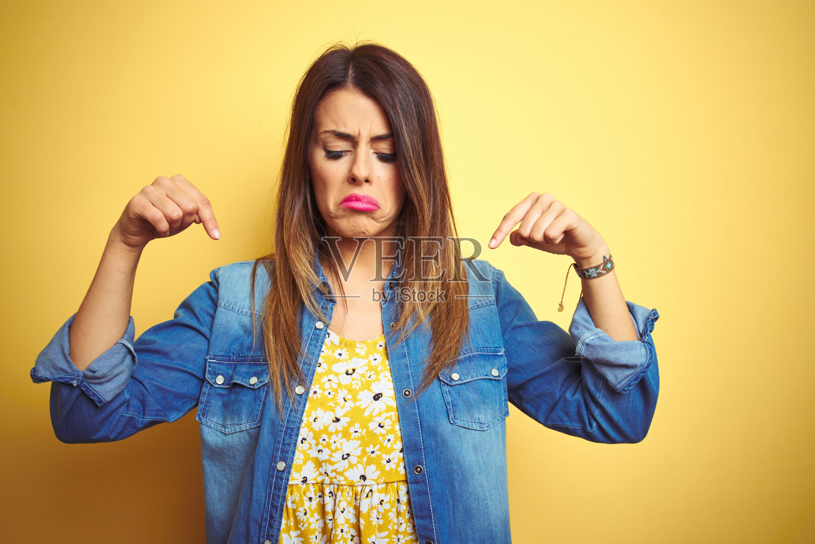 年轻漂亮的女人站在黄色背景前，指着下方，看起来悲伤和沮丧照片摄影图片