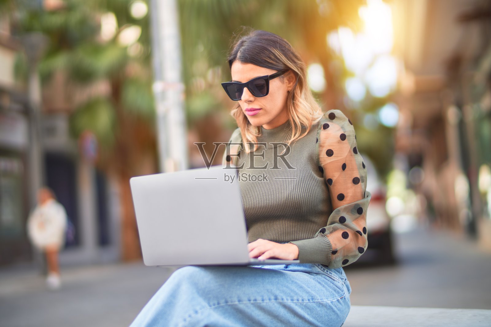 年轻漂亮的女人戴着太阳镜，微笑快乐和自信。坐在长椅上使用笔记本电脑照片摄影图片