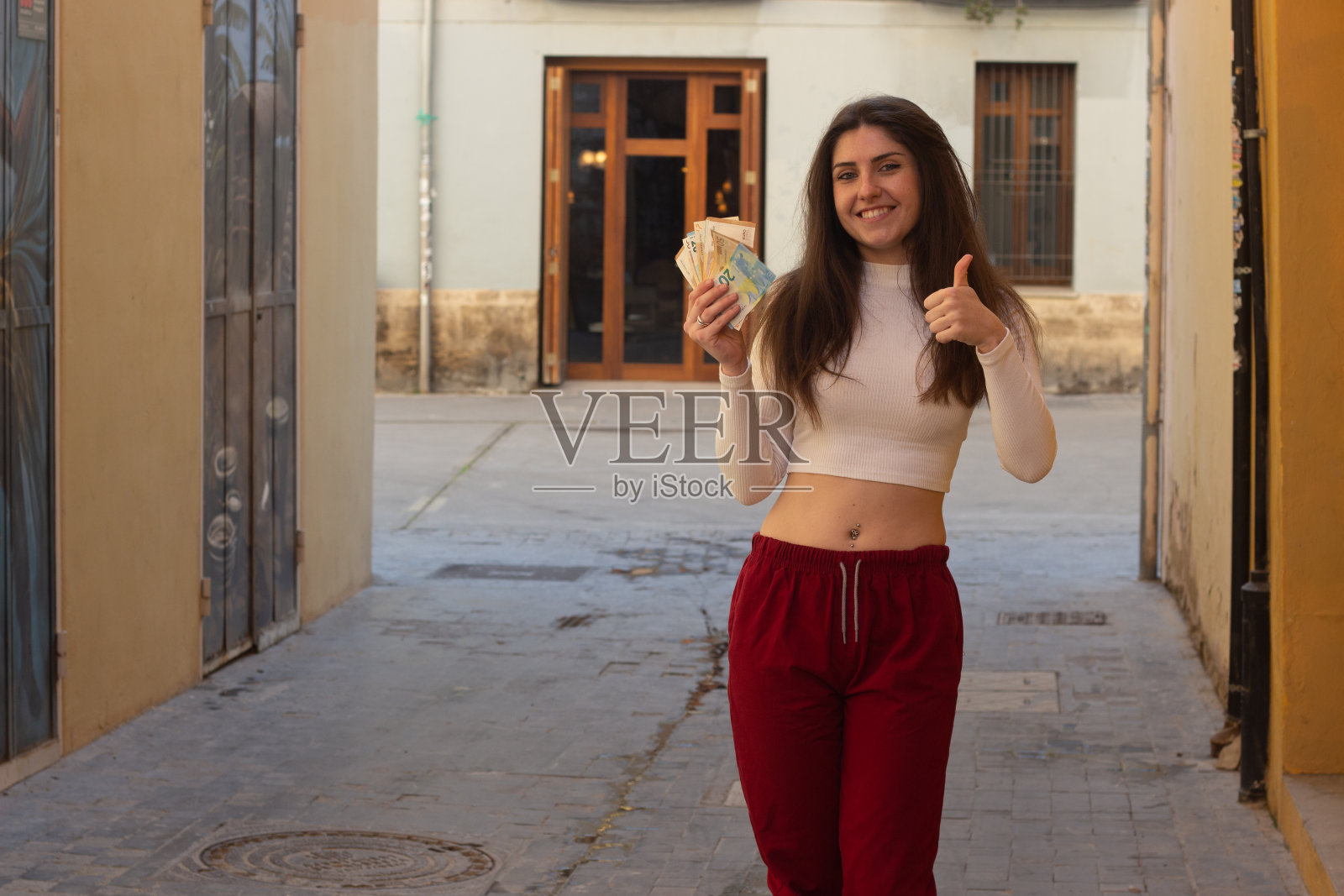 画像中迷人的白种年轻女子模特用拇指向上的手势与欧元钞票，在城市里，白色的上衣和红色的裤子，黑暗的空气。将你的文本放在拷贝空间。照片摄影图片