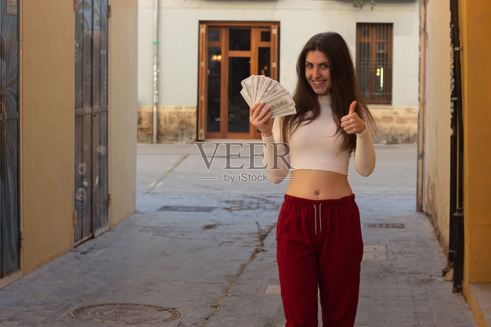 迷人的年轻女子用美钞翘起拇指的手势，在城市里，白色的上衣和红色的裤子，黑暗的空气中。将你的文本放在拷贝空间。照片摄影图片