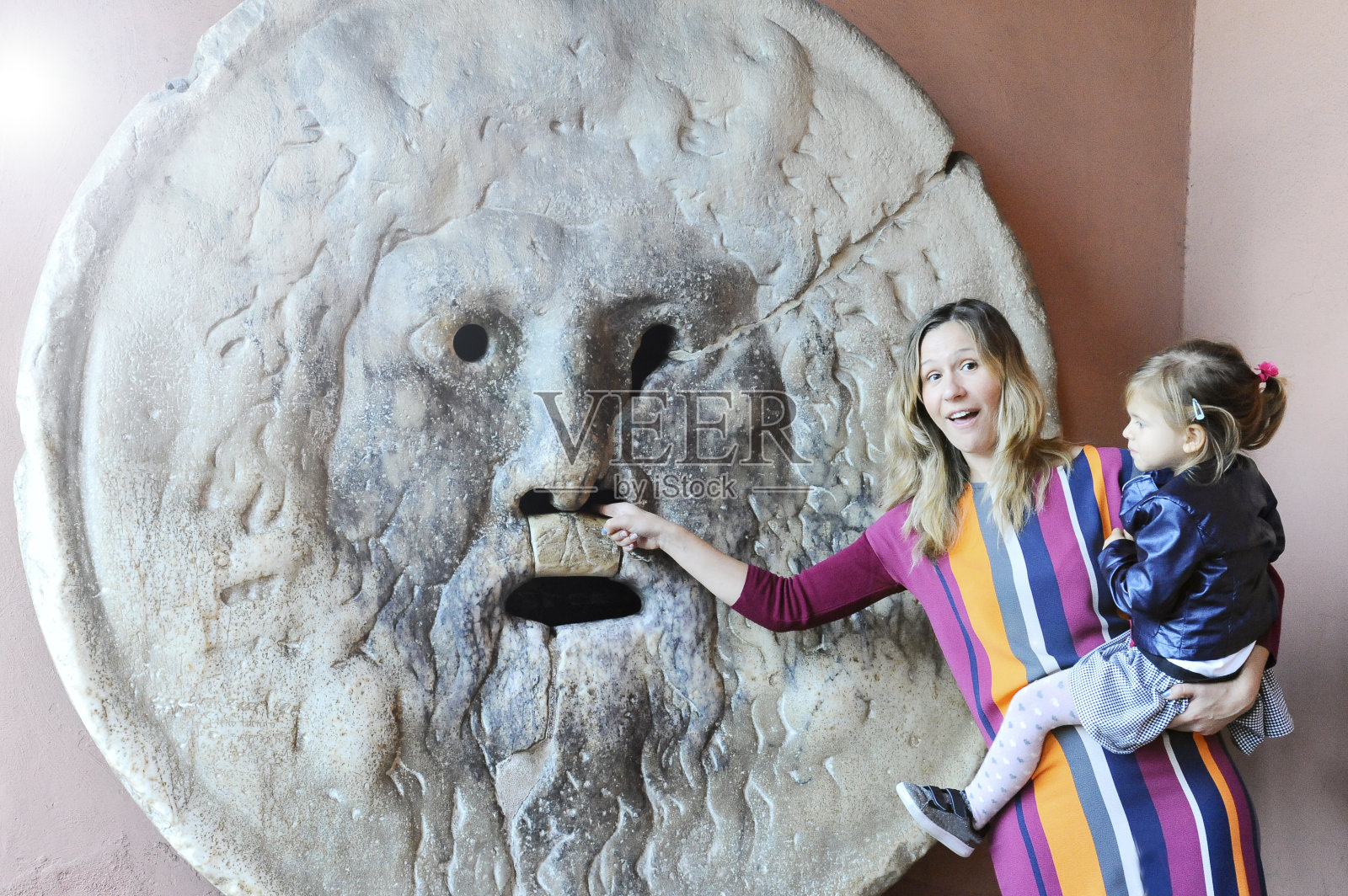 在真理之口游玩的微笑的年轻母女游客。母亲穿着彩色连衣裙，女儿穿着蓝色皮夹克。有趣的场景。意大利罗马照片摄影图片