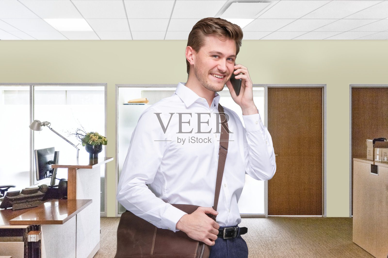 男性员工站在办公室的小隔间前，穿着邮差包，拿着包，使用智能手机照片摄影图片