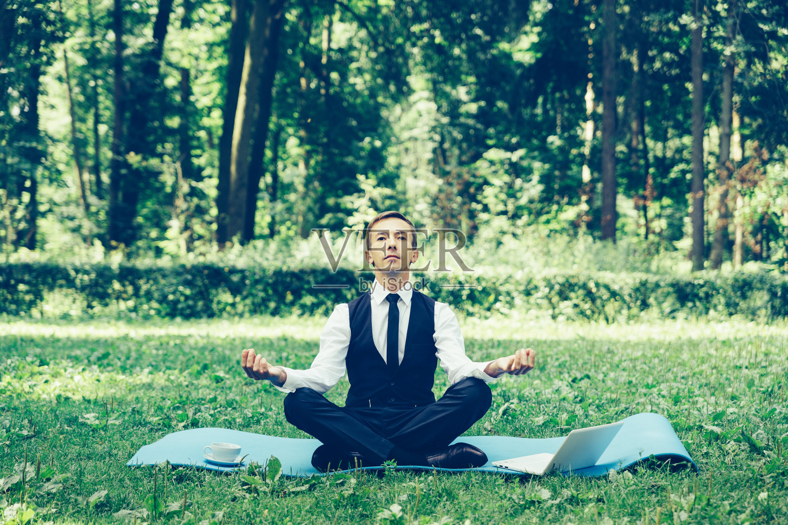 商人在公园里以莲花姿势放松，一手拿着笔记本电脑，一手拿着茶杯，闭着眼睛，面容平静，找到了平衡照片摄影图片